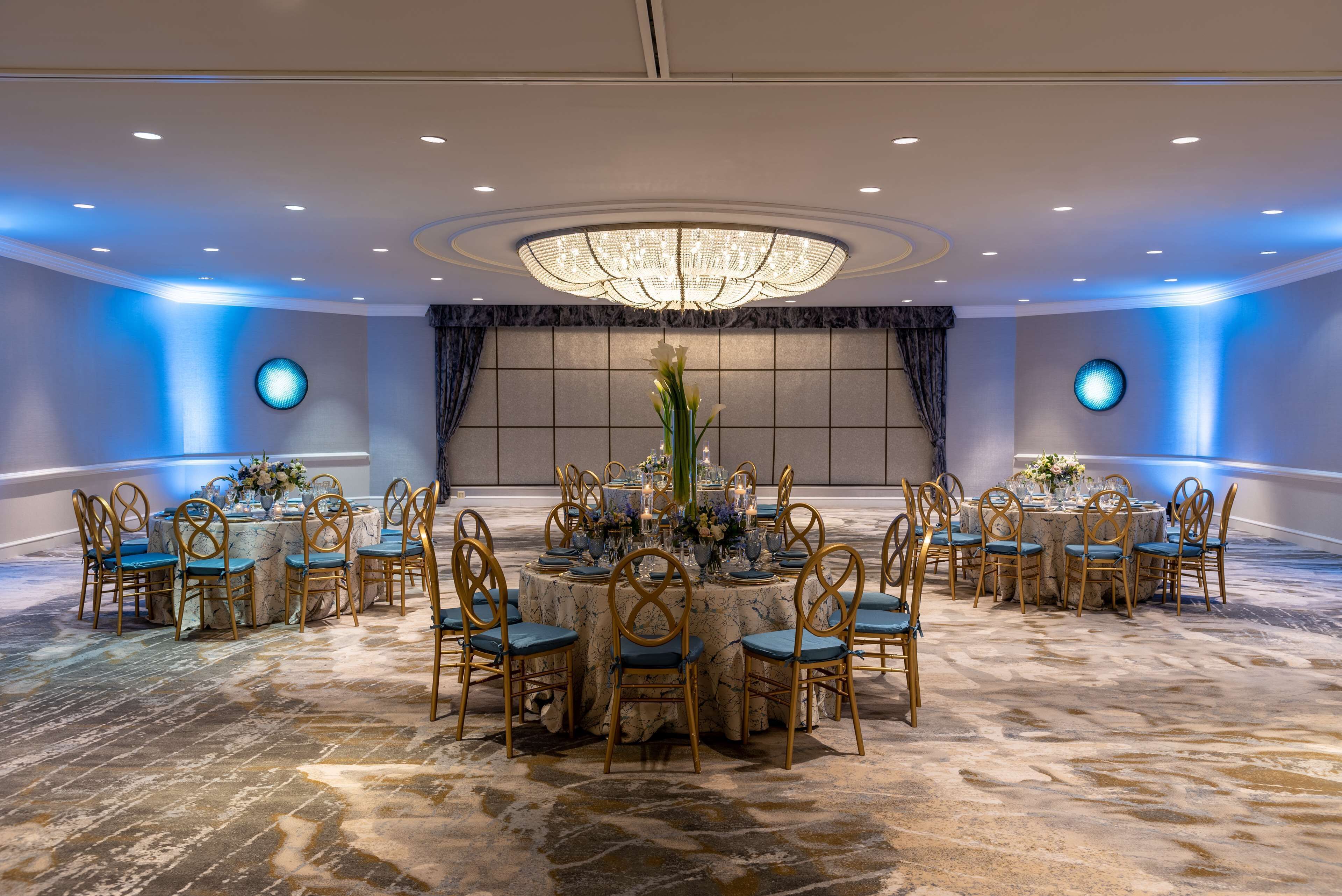 Un amplio salón de banquetes con mesas redondas y sillas, un candelabro central e iluminación ambiental azul.