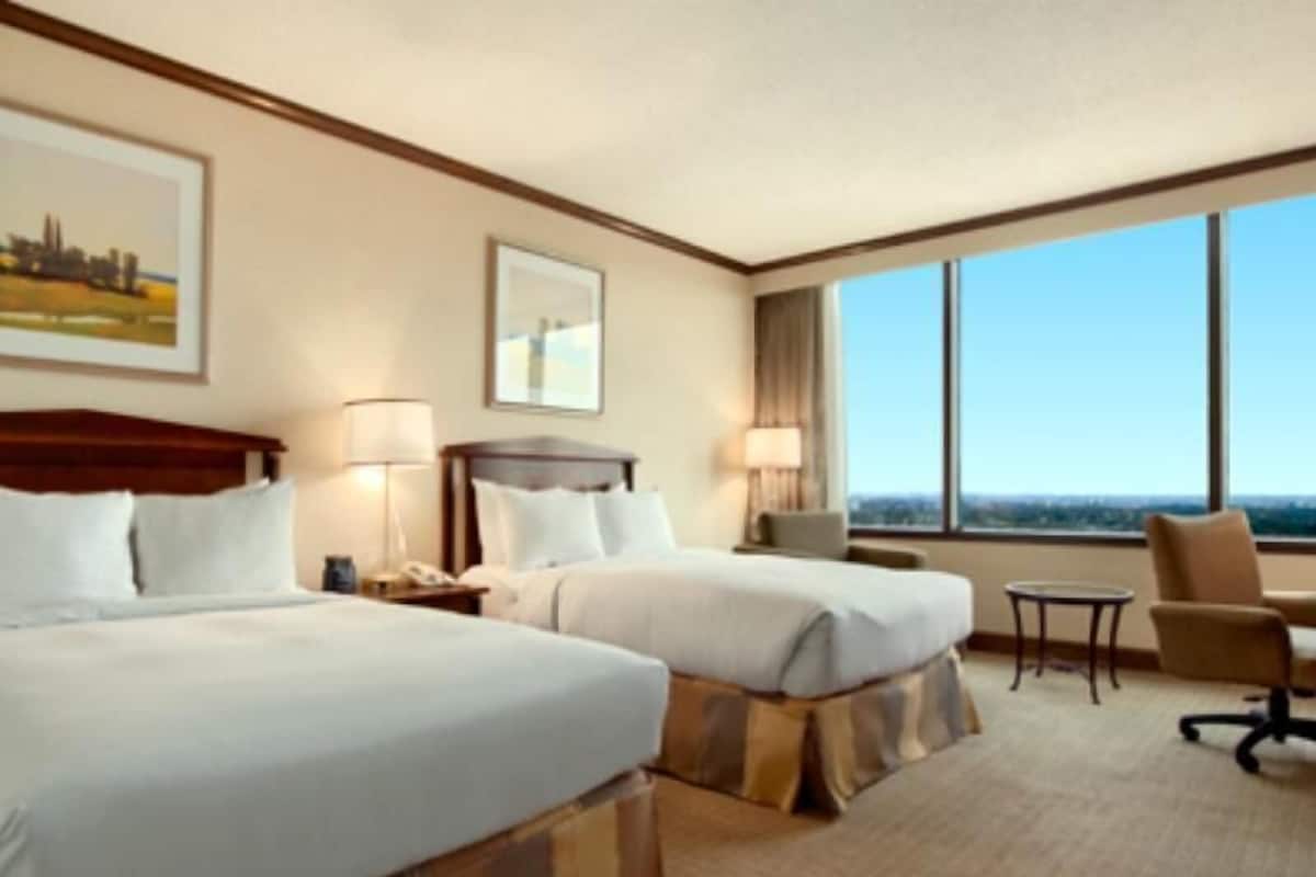 Ein Hotelzimmer mit zwei Betten, einem Schreibtisch, einem Stuhl und einem großen Fenster mit Blick auf die Stadt.