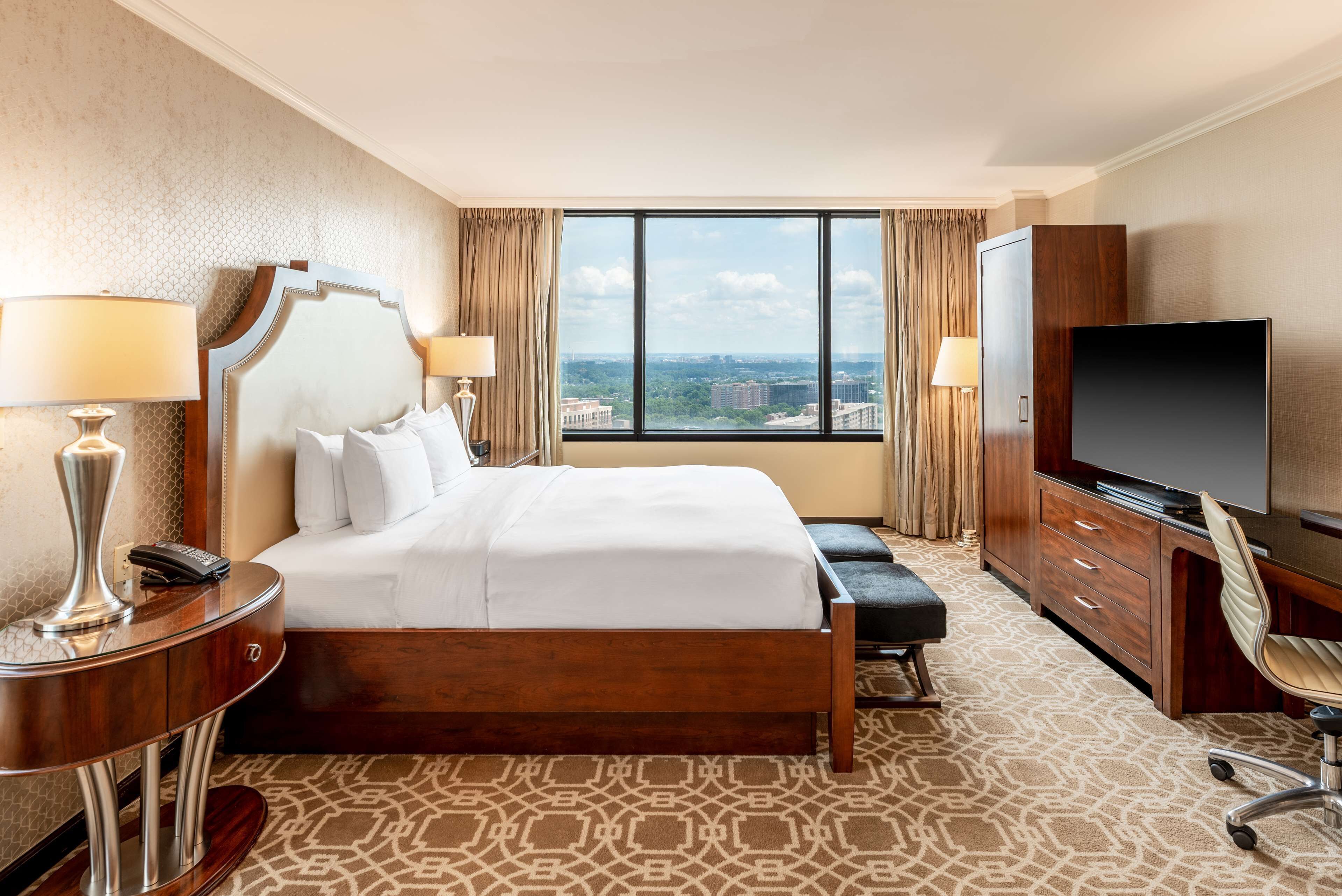 A hotel room with a large bed, a desk with a television, and a view of the city through a large window.