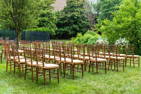 Rows of wooden chairs with cushions arranged in a grassy outdoor area, surrounded by trees and a fence.