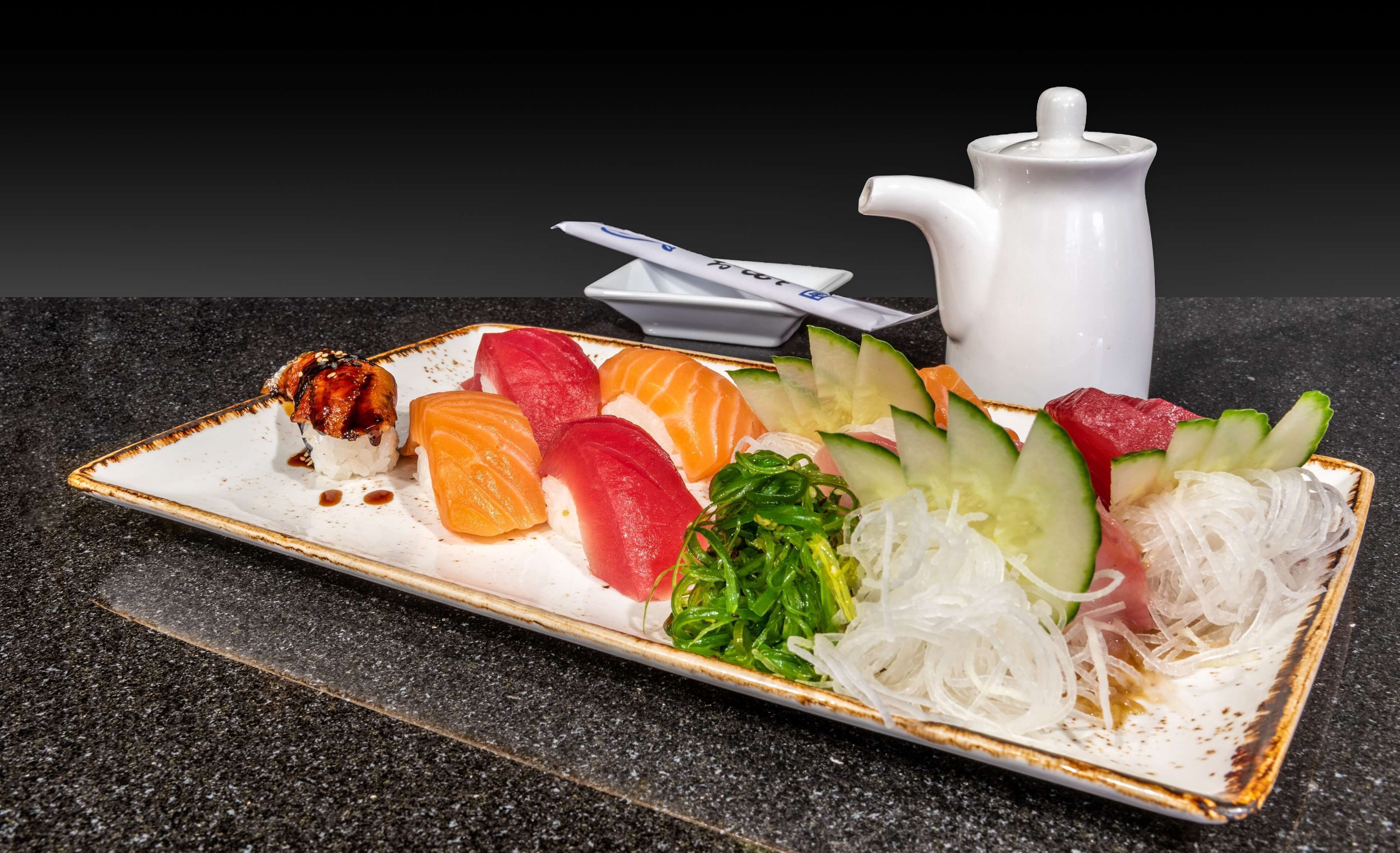 A sushi platter with various types of fish, vegetables, and a white ceramic pitcher on a rectangular plate.