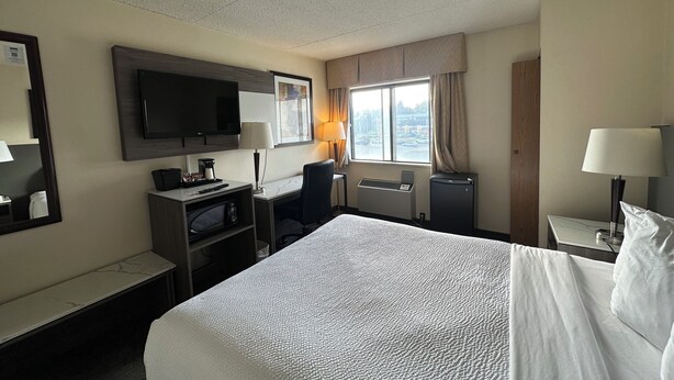 Ein Hotelzimmer mit einem großen Bett, einem Schreibtisch mit Computer, einem Fernseher und einem Fenster mit Vorhängen.