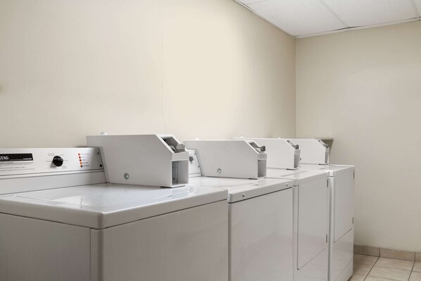 Une buanderie équipée de quatre machines à laver et de quatre sèche-linge à chargement frontal, tous deux de couleur blanche, d’un panneau de commande et d’un sol carrelé.