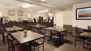 Un espace repas avec plusieurs tables et chaises, un coin cuisine équipé d’appareils électroménagers et des tableaux encadrés accrochés aux murs.
