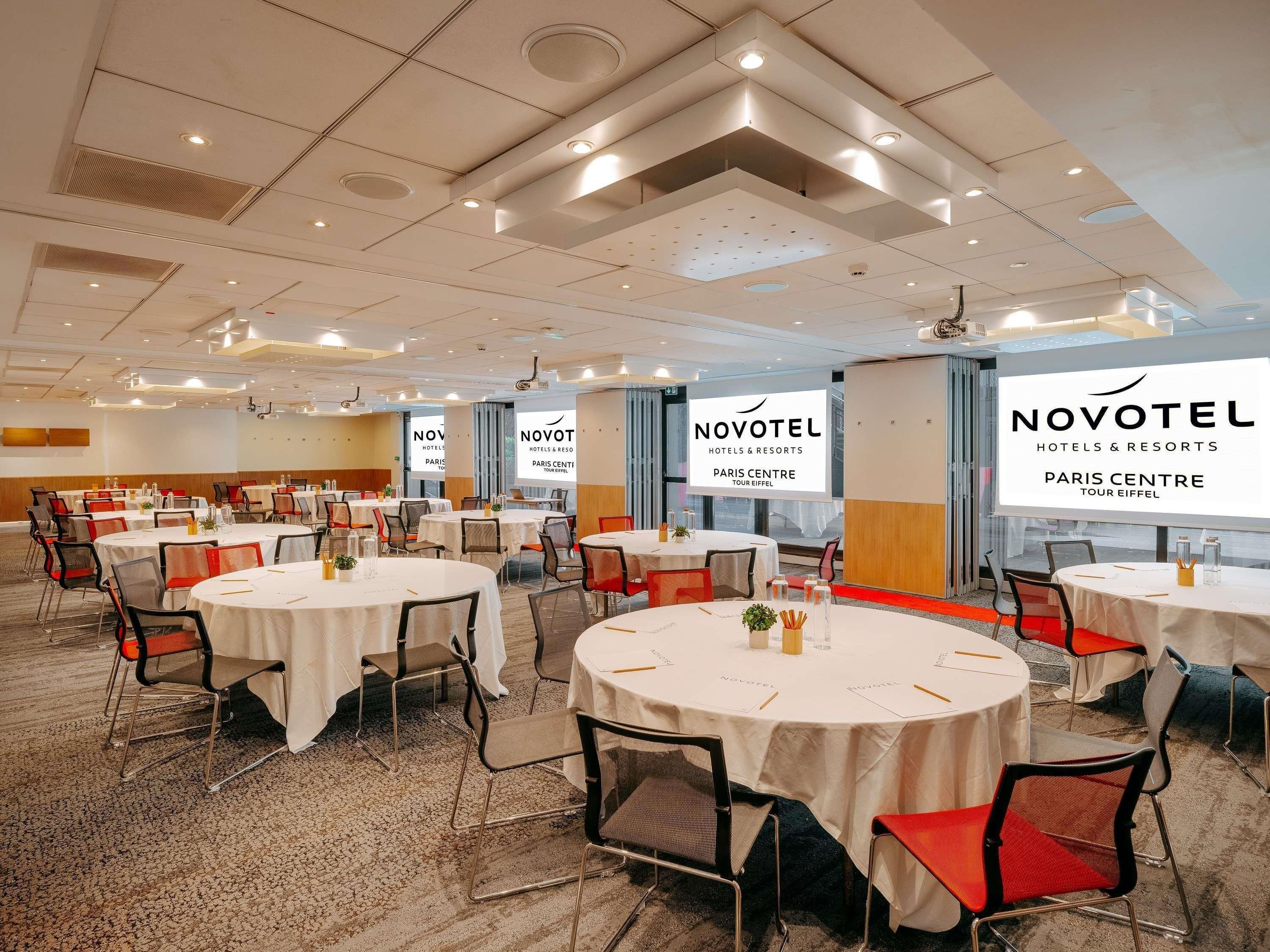 Ein Konferenzraum in einem Hotel mit runden Tischen, Stühlen und einer großen Leinwand, auf der das Novotel-Logo zu sehen ist.