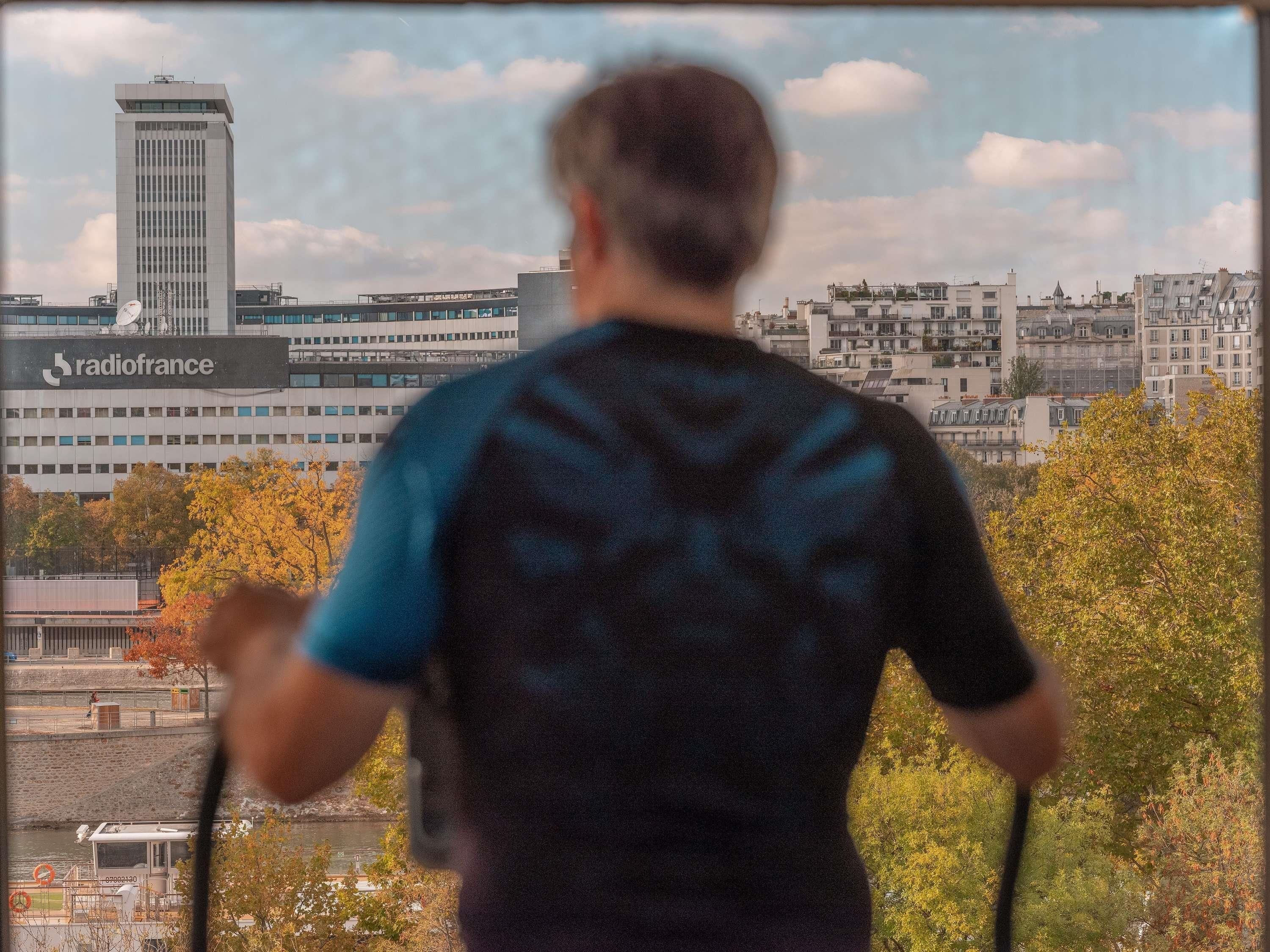 Une personne se tient debout, face à un panorama urbain, qui comprend un grand immeuble sur lequel on peut lire l’inscription « radiofrance ».