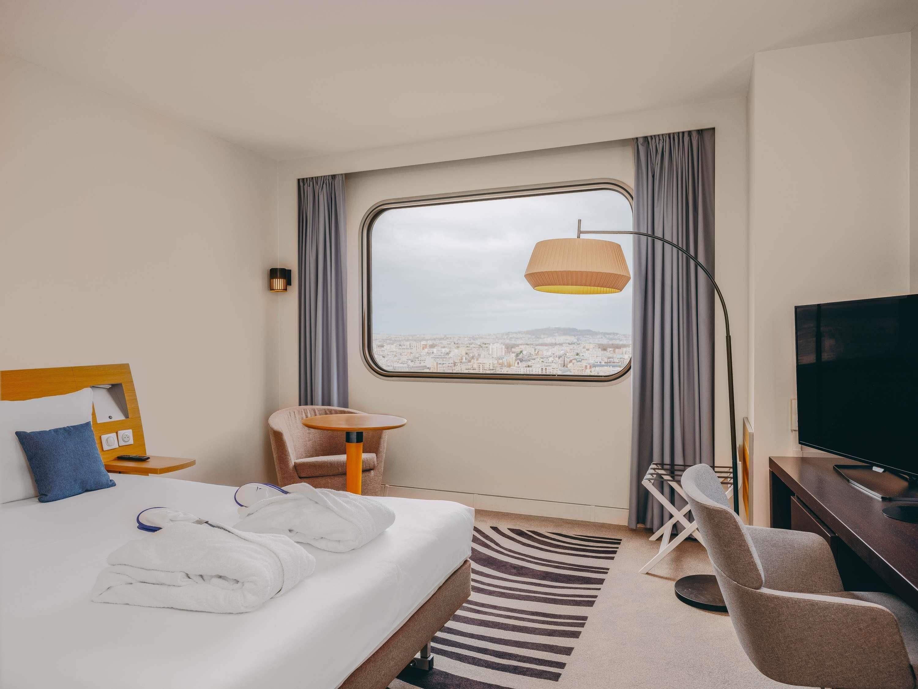 A hotel room with a bed, a desk, a chair, a TV, and a window with a city view.