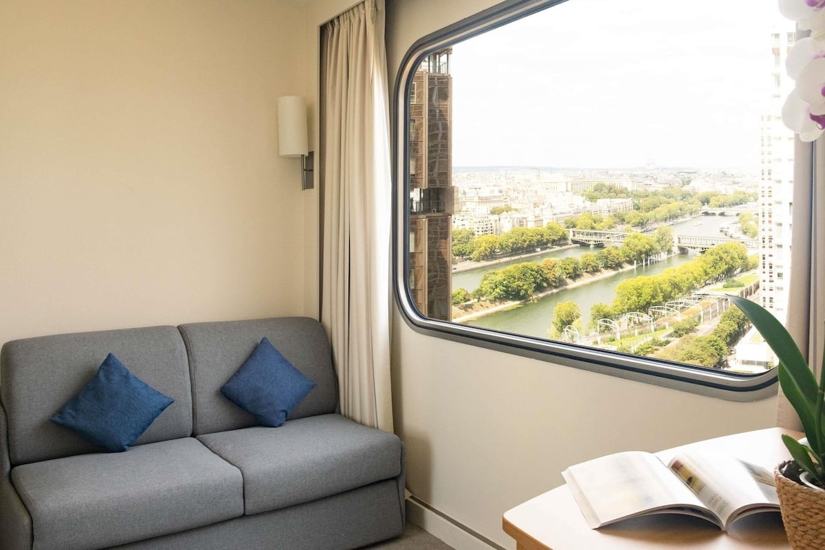 Una habitación con un sofá gris, dos cojines azules, un ventanal con vistas a un paisaje urbano y una mesa con un libro abierto y una planta en maceta.