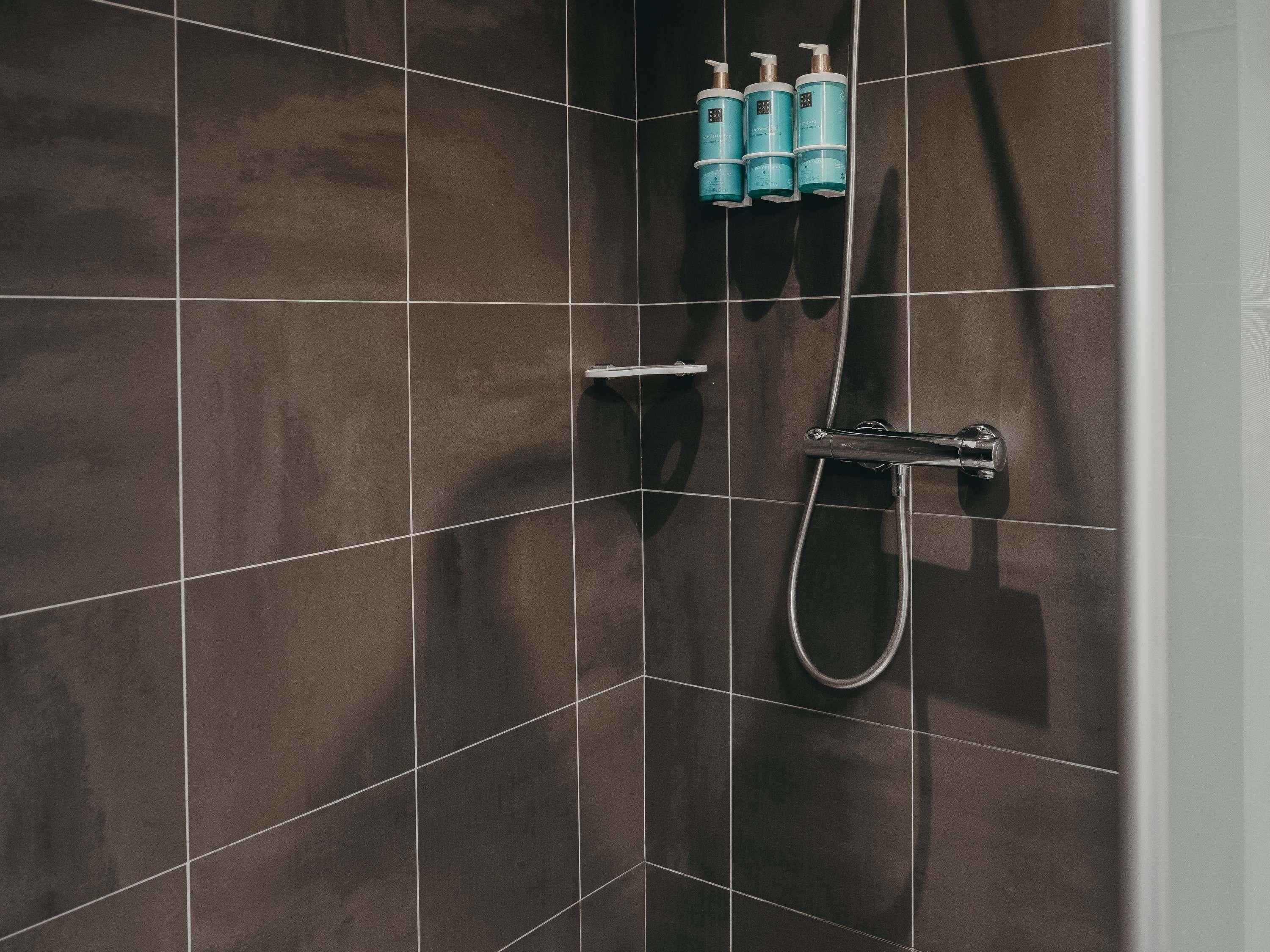A tiled shower wall with a handheld showerhead and three bottles of shampoo.