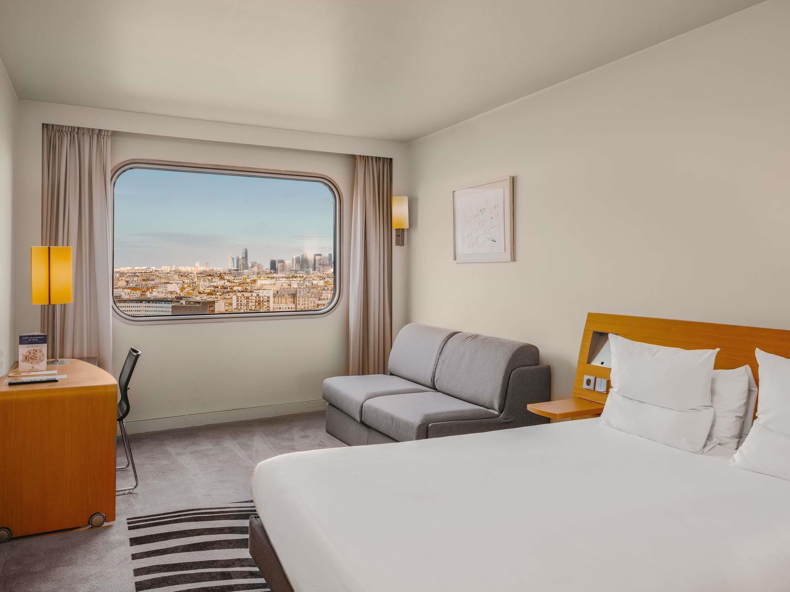 Ein Hotelzimmer mit Bett, Sofa, Schreibtisch mit Lampe, Stuhl und Fenster mit Stadtblick.
