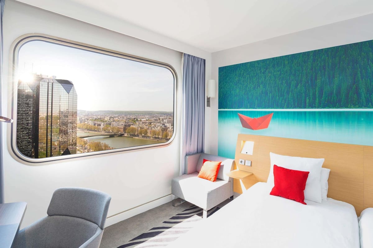 Habitación de hotel con un amplio ventanal que ofrece vistas a la ciudad, una cama con ropa blanca y almohadas rojas, un sillón gris y una obra de arte moderna en la pared.