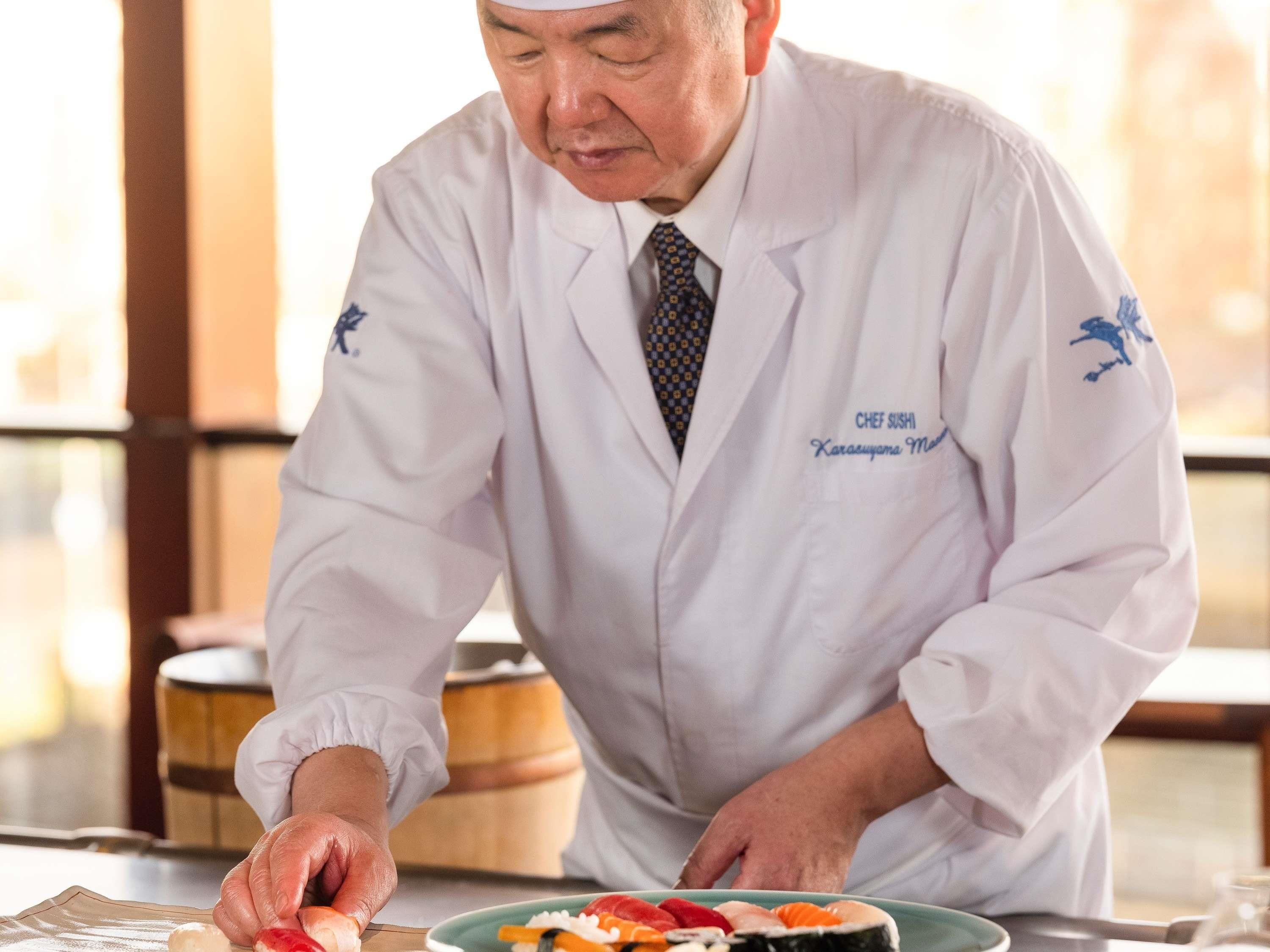 A person in a white lab coat with 'CHEF SUSHI' and 'Karasuyama Michi' embroidered on it, preparing sushi at a table.