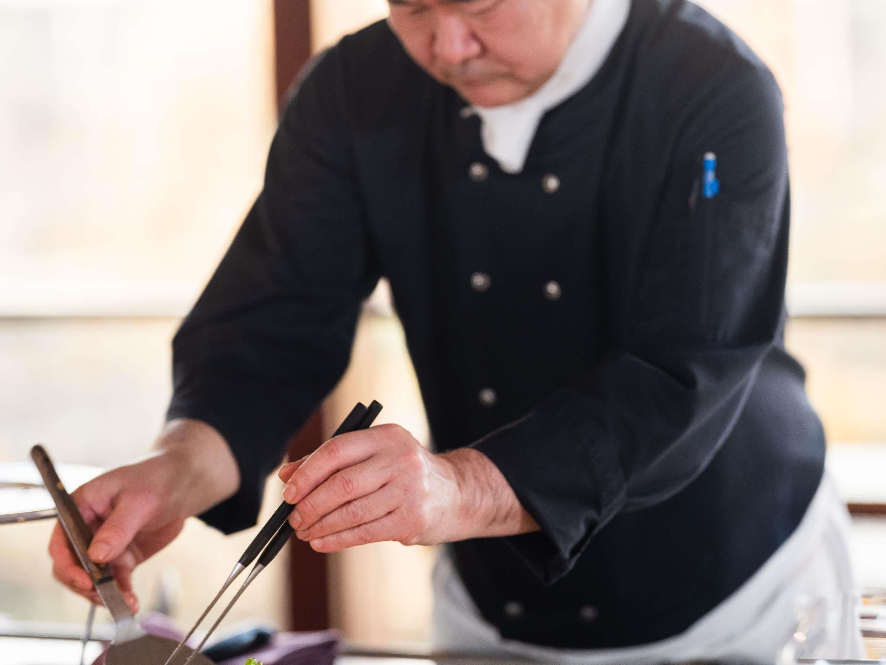 A person in a black chef's coat using chopsticks and a knife on a table.