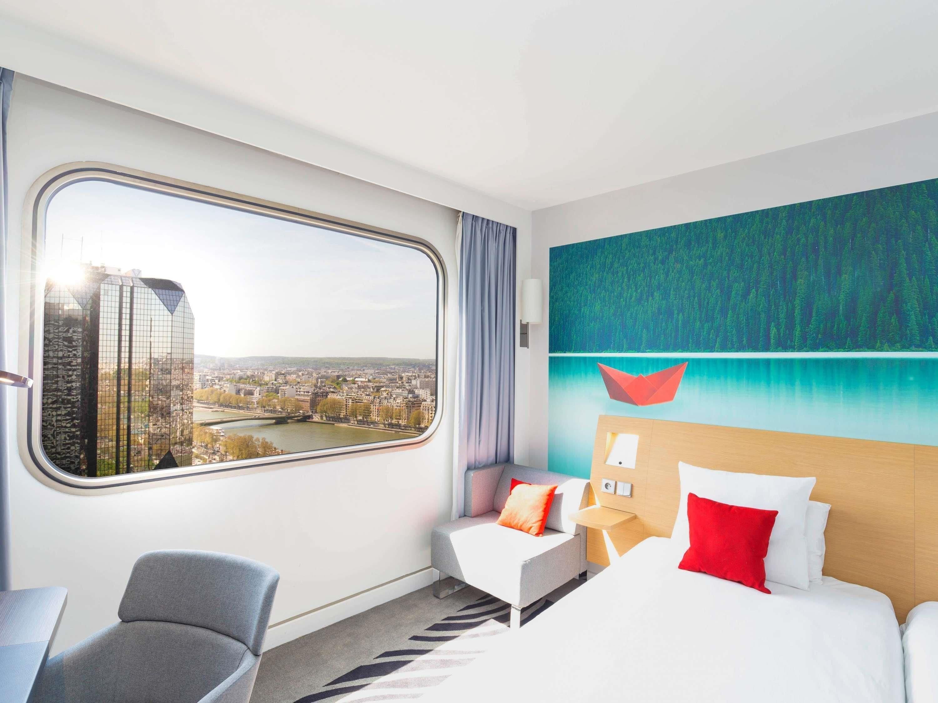 Une chambre d’hôtel dotée d’une grande fenêtre donnant sur la ville, d’un lit avec une literie blanche et des oreillers rouges, d’un fauteuil gris et d’une œuvre d’art moderne accrochée au mur.