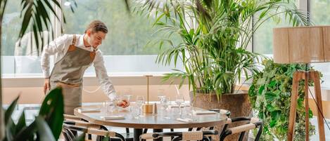 A person setting up a table with wine glasses in a room with large windows, potted plants, and a wooden lamp.