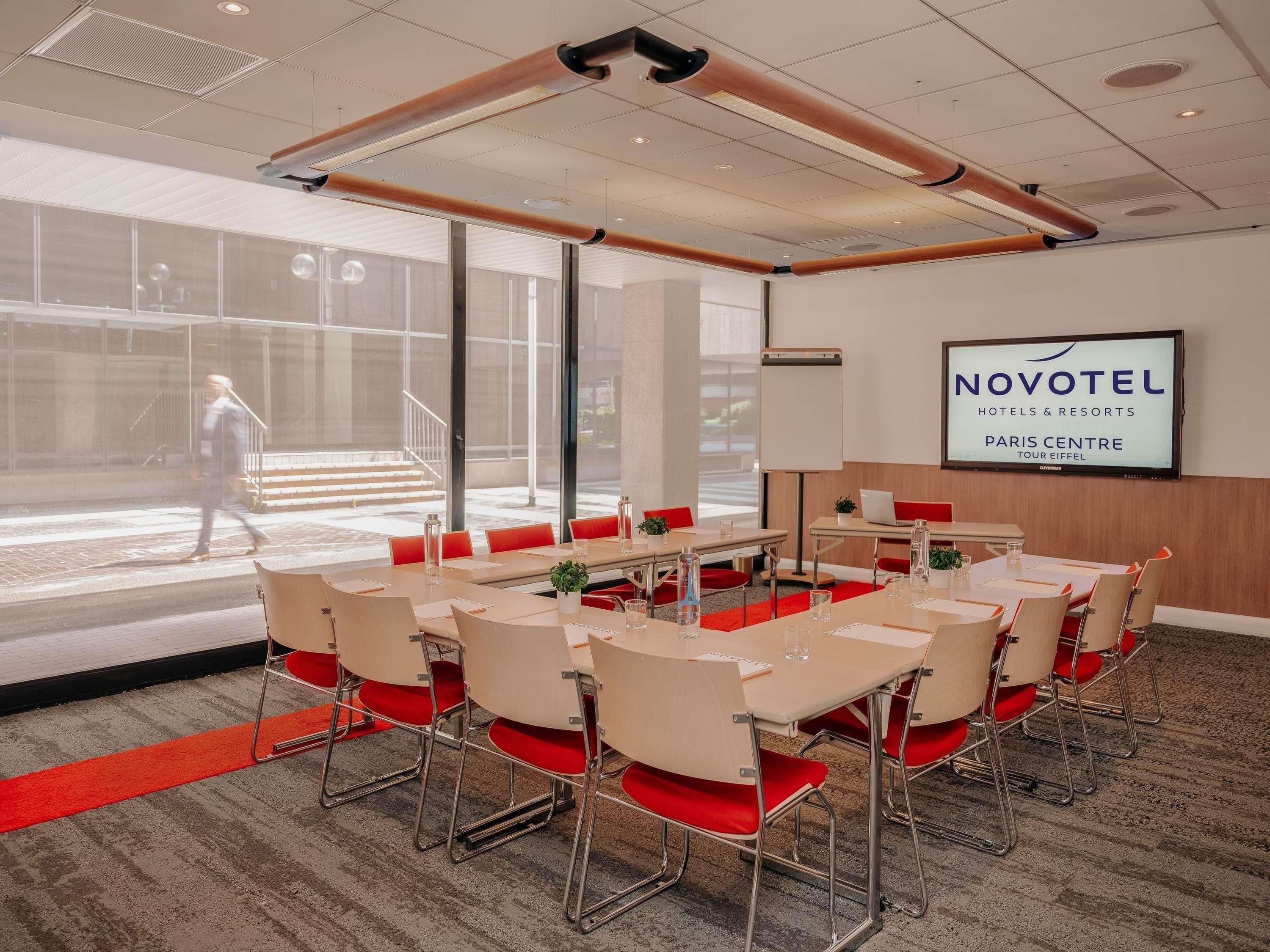 Un salón de conferencias con una gran mesa, sillas rojas, una pizarra y una pantalla de televisión mostrando 'NOVOTEL HOTELS & RESORTS PARIS CENTRE TOUR EIFFEL'.