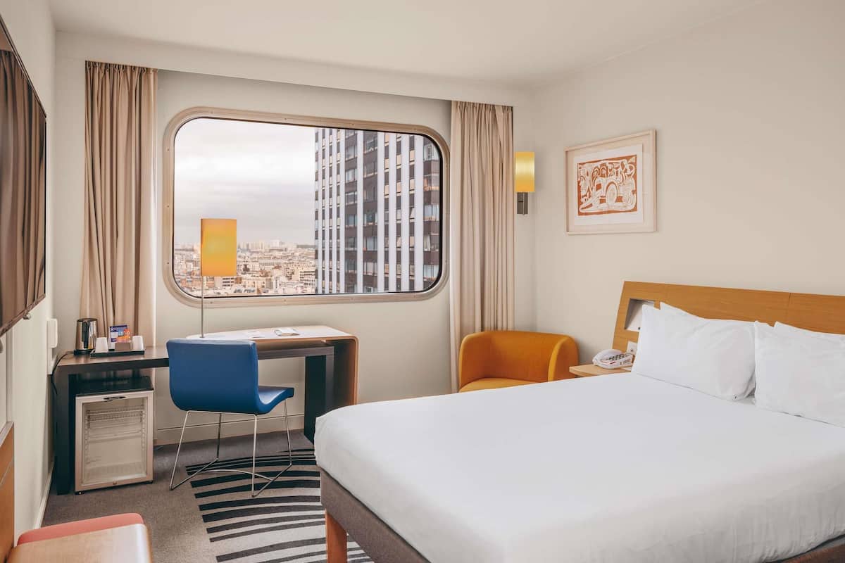 Habitación de hotel con cama, escritorio, silla, refrigerador, ventana con vista a la ciudad y una lámpara de pared.