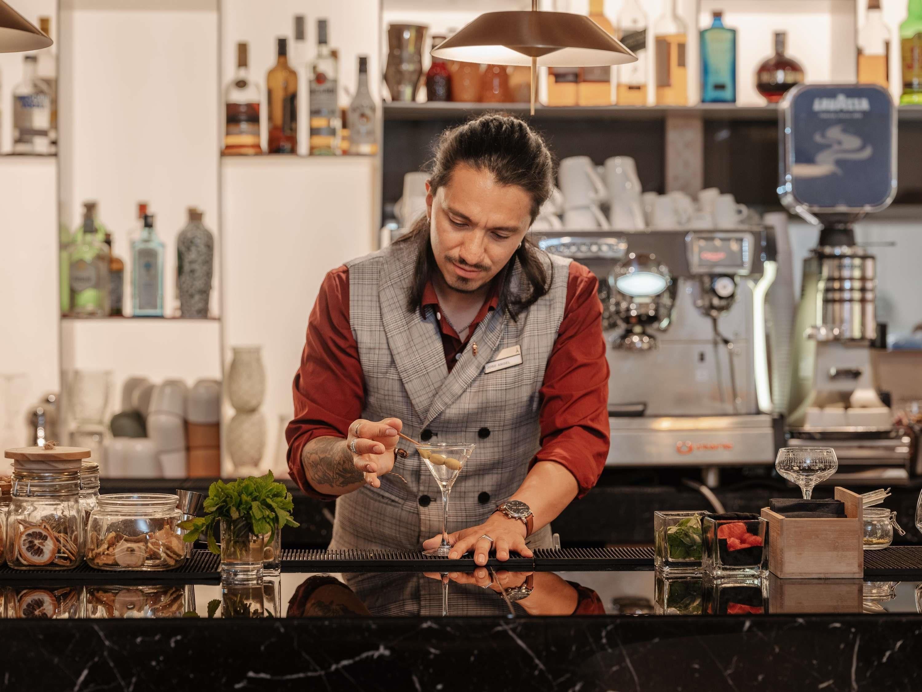 A bartender at a bar with a coffee machine and various bottles in the background.