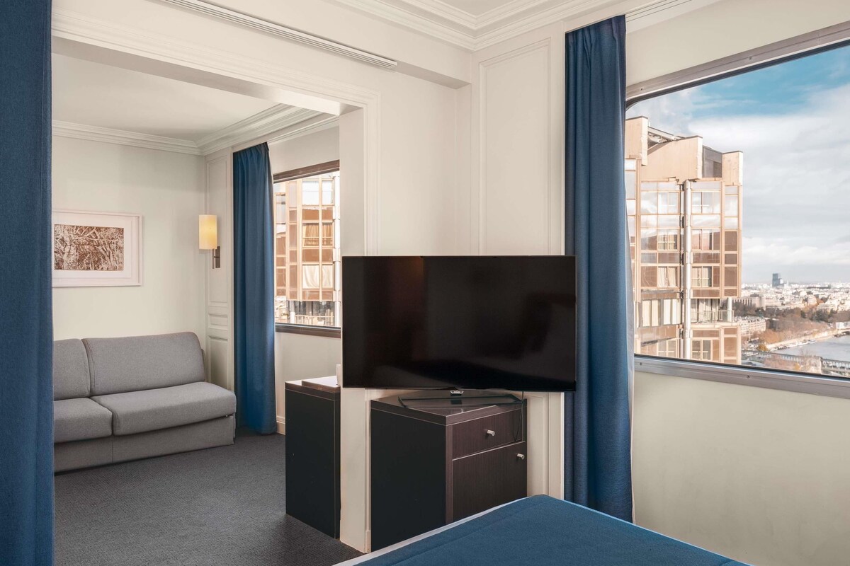 Habitación de hotel con cama, televisión, escritorio, sofá y ventana con vista a la ciudad.