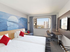 A hotel room with two beds, a desk, a chair, a TV, a large window with a city view, and a mural on the wall.