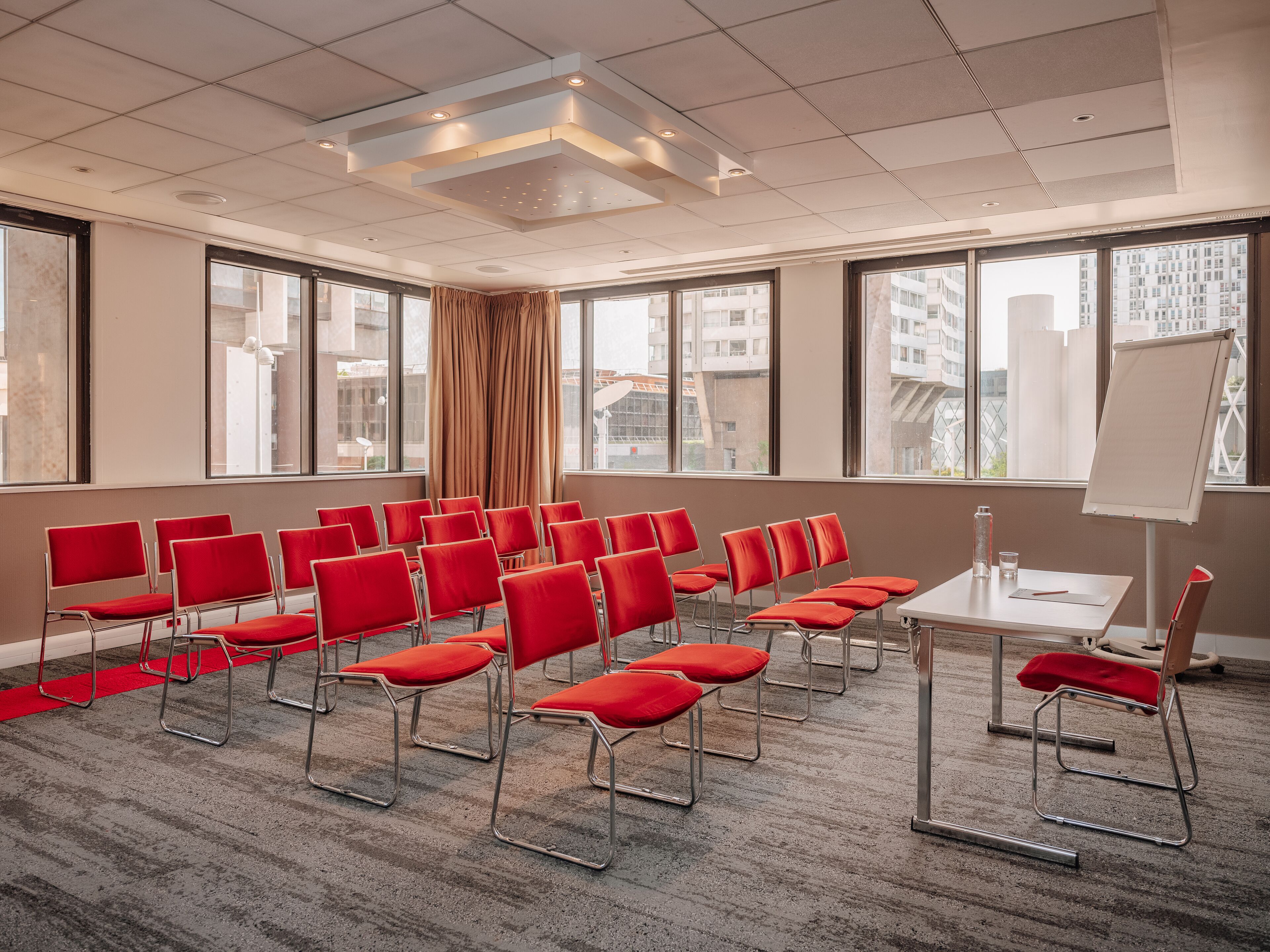Sala de conferências com fileiras de cadeiras vermelhas, um quadro branco e amplas janelas com vista para a cidade.