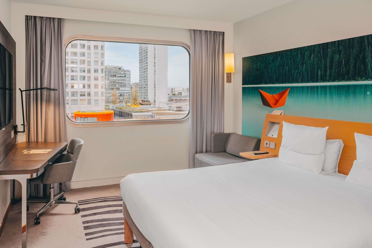 Habitación de hotel con cama, escritorio, silla, televisor, ventana con vista a la ciudad y un cuadro en la pared.