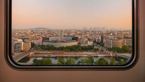 A panoramic view of a cityscape through a window, featuring a prominent building with a circular structure and a river with a bridge. - Novotel Paris Centre Tour Eiffel (Paris)