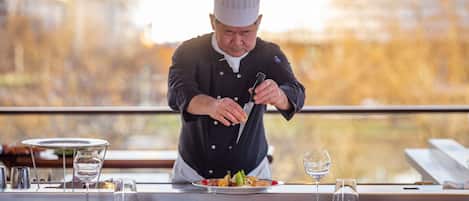 Un chef preparando comida en una mesa de restaurante con vista a un cuerpo de agua.