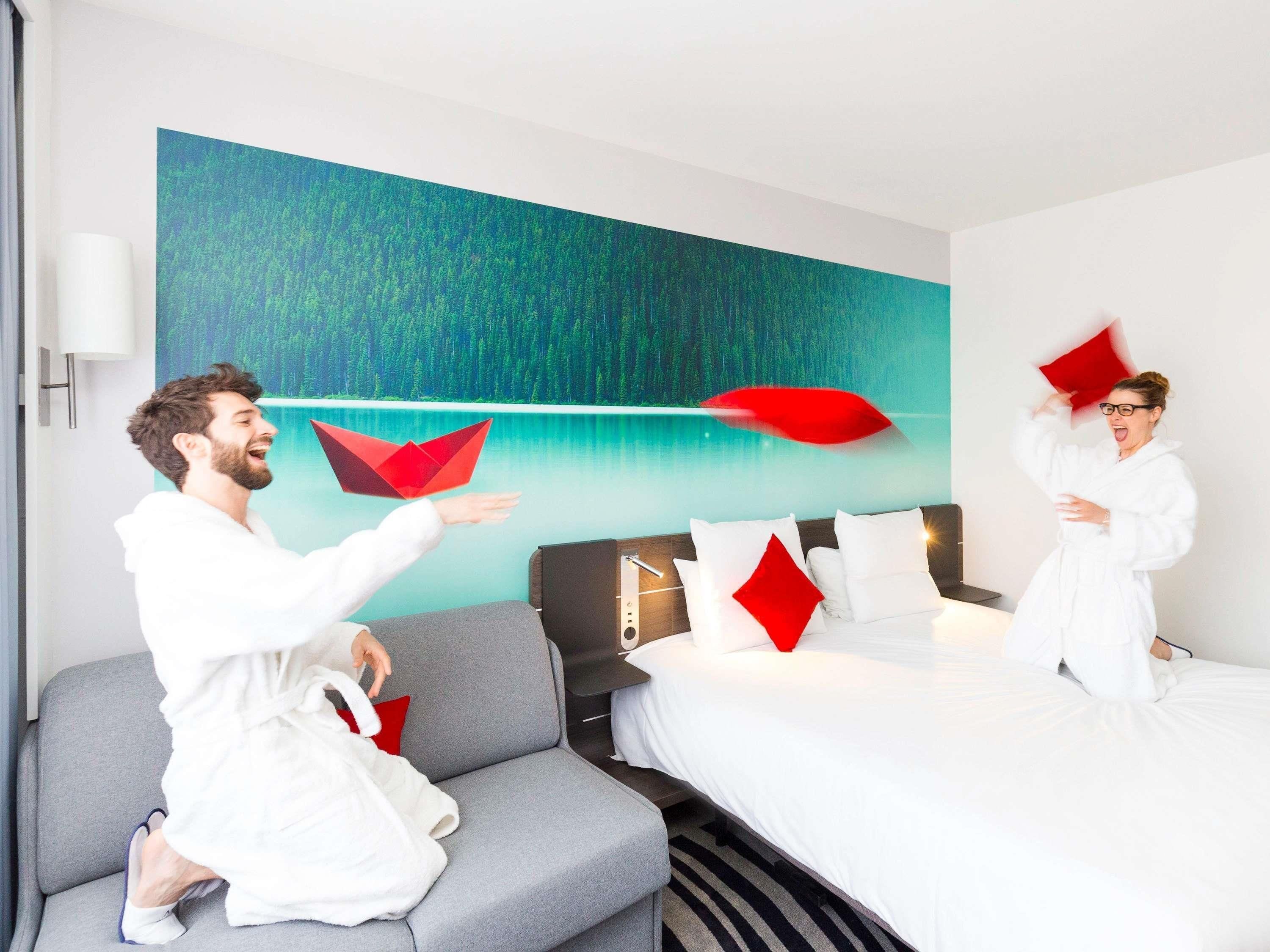 Une chambre d’hôtel avec un lit, un canapé gris et un tableau représentant une forêt accroché au mur.