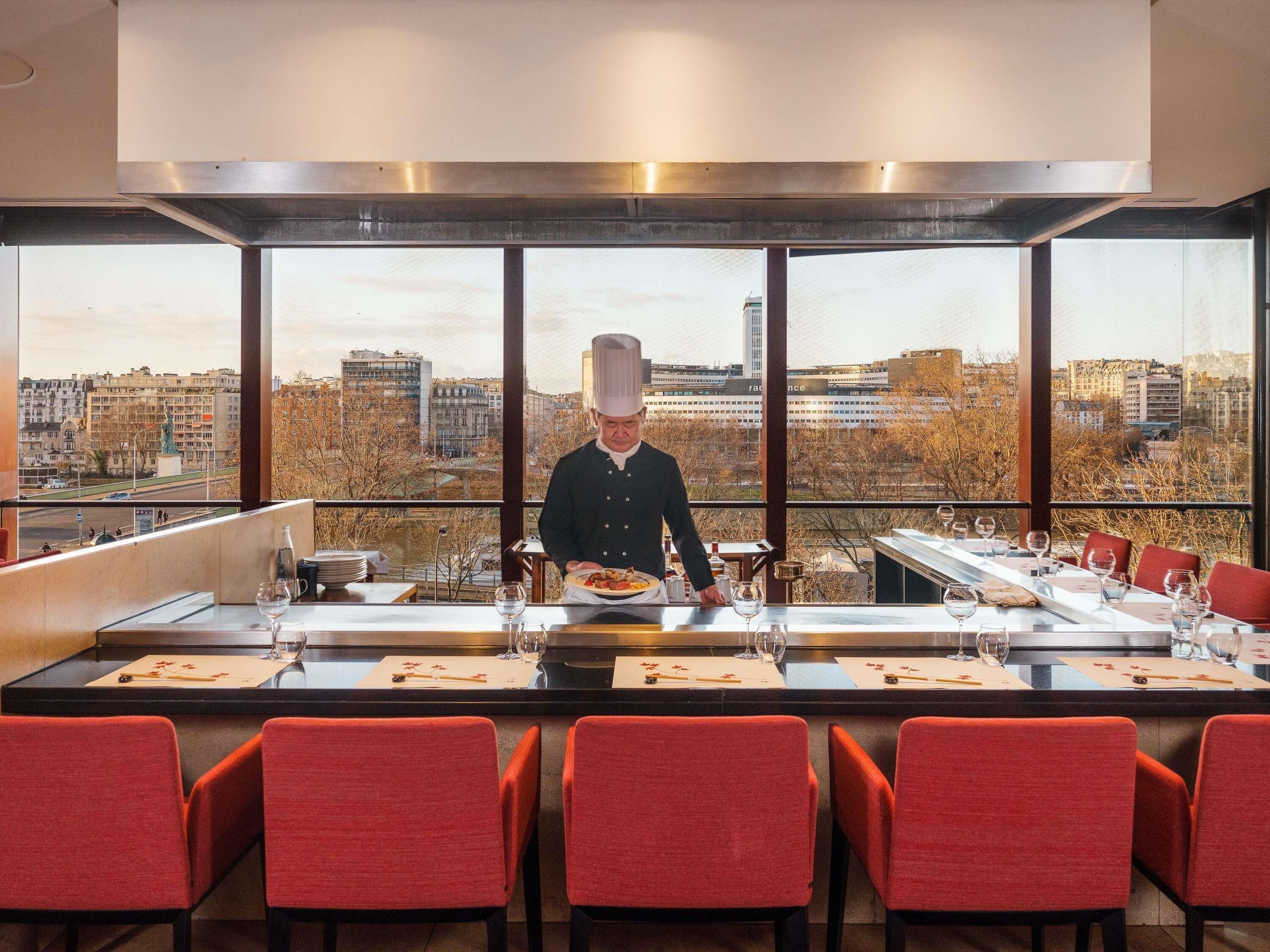 Een kok bereidt eten aan een tafel met uitzicht over de stad, rode stoelen en een modern interieur.