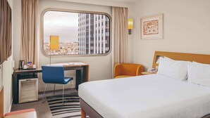 Een hotelkamer met een bed, een bureau, een stoel, een kleine koelkast en een raam met uitzicht op de stad.