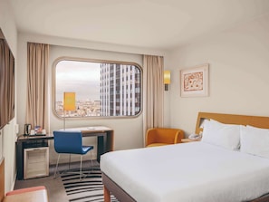 Habitación de hotel con cama, escritorio, silla, refrigerador pequeño y ventana con vista a la ciudad.