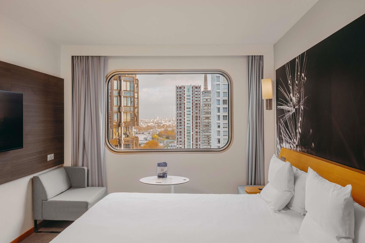 Habitación de hotel con una cama grande, un televisor en la pared, una mesita y vistas a edificios altos.