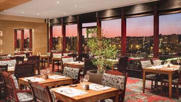 Un restaurant avec des tables dressées pour le service, de grandes fenêtres offrant une vue sur la ville et un arrangement floral en guise de décoration centrale.