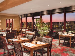 Een restaurant met gedekte tafels, grote ramen met uitzicht op de stad en een planten decoratie.
