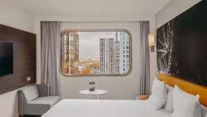 A hotel room with a large bed, a TV mounted on the wall, a small table, and a view of tall buildings. - Novotel Paris Centre Tour Eiffel (Paris)