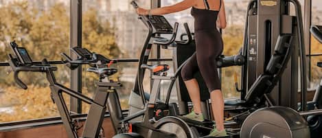 A gym with a treadmill, weight machines, and a view of buildings through large windows.
