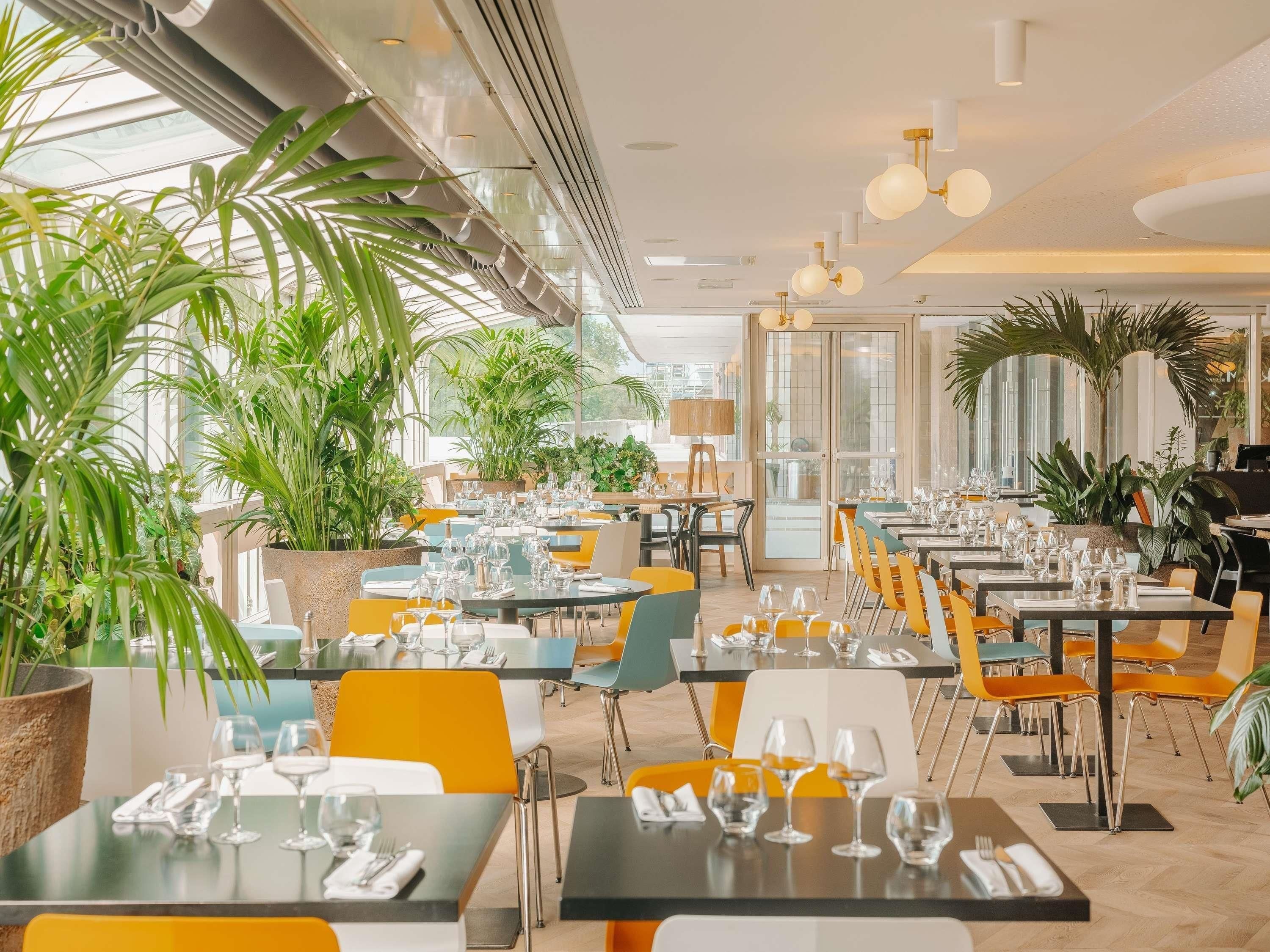Restaurante com plantas internas, mesas dispostas com copos e talheres, e design de interiores moderno.