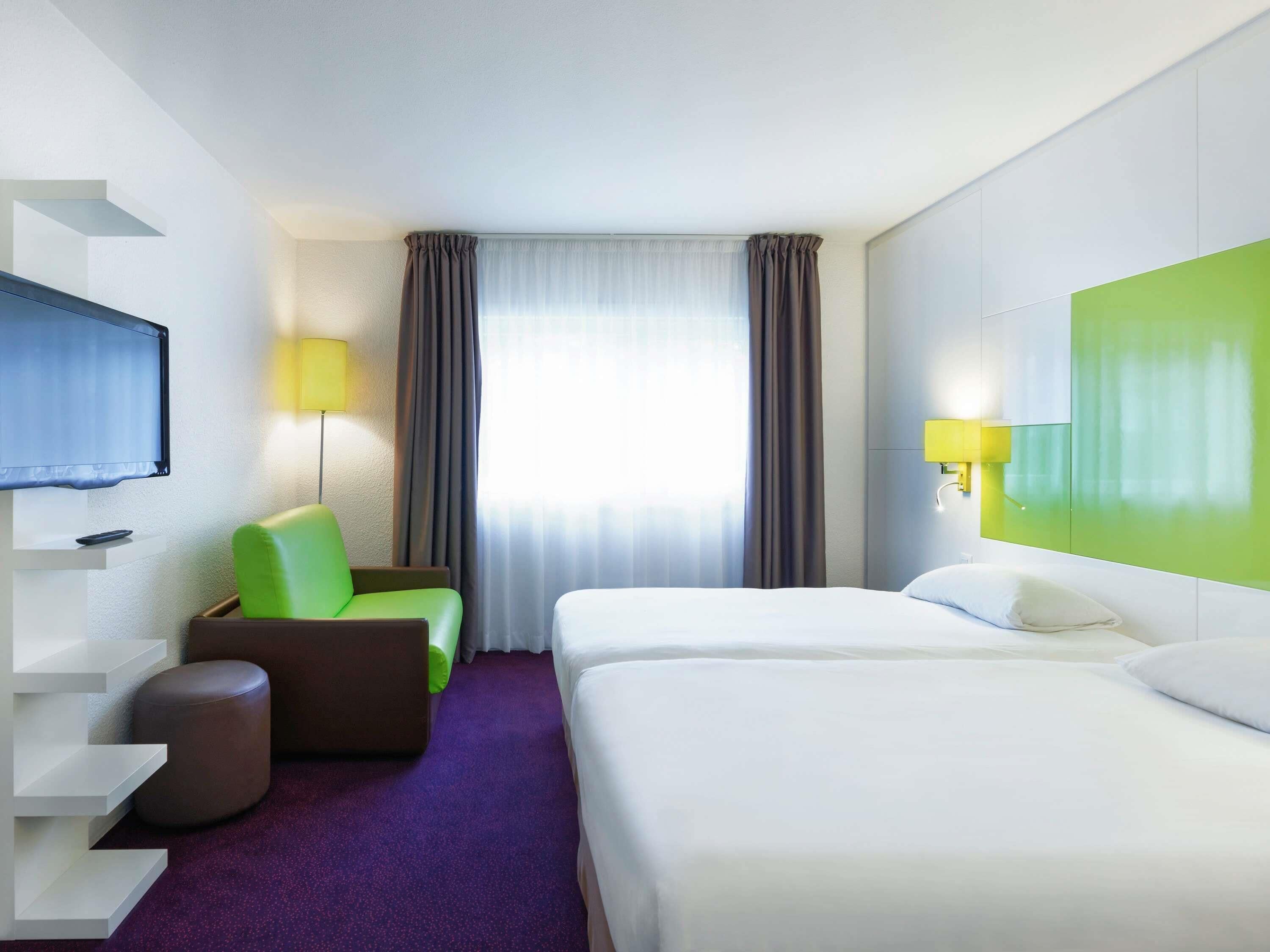 Une chambre d’hôtel avec un lit, une chaise, une télévision, une lampe et une fenêtre avec des rideaux.