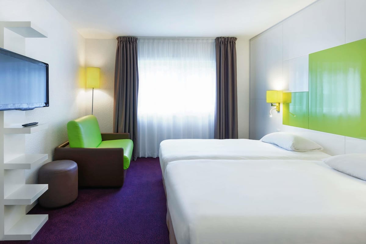 A hotel room with a bed, a chair, a TV, a lamp, and a window with curtains.
