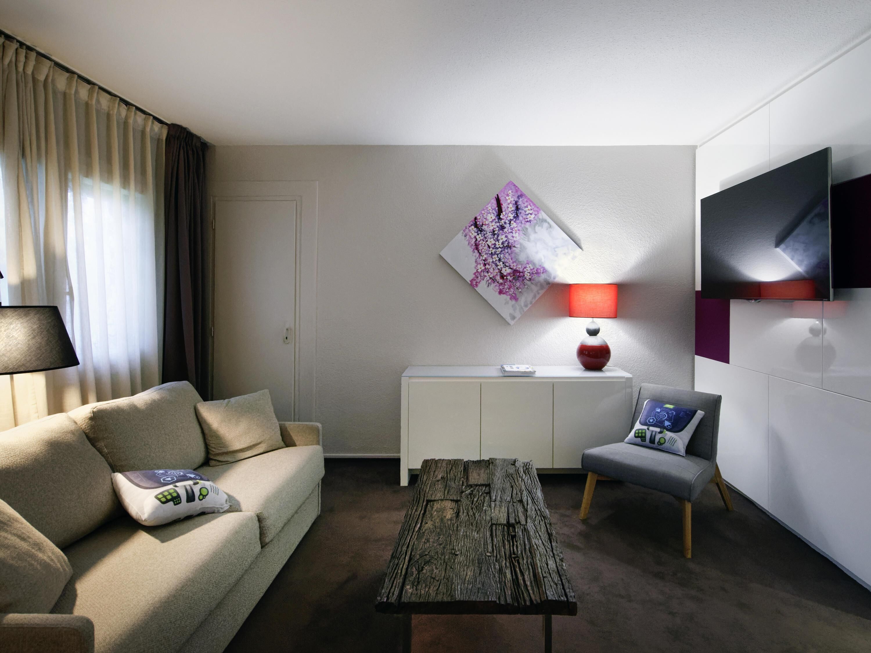 Una habitación de hotel moderna con sofá, una mesa de centro, televisor, un cuadro y una lámpara.