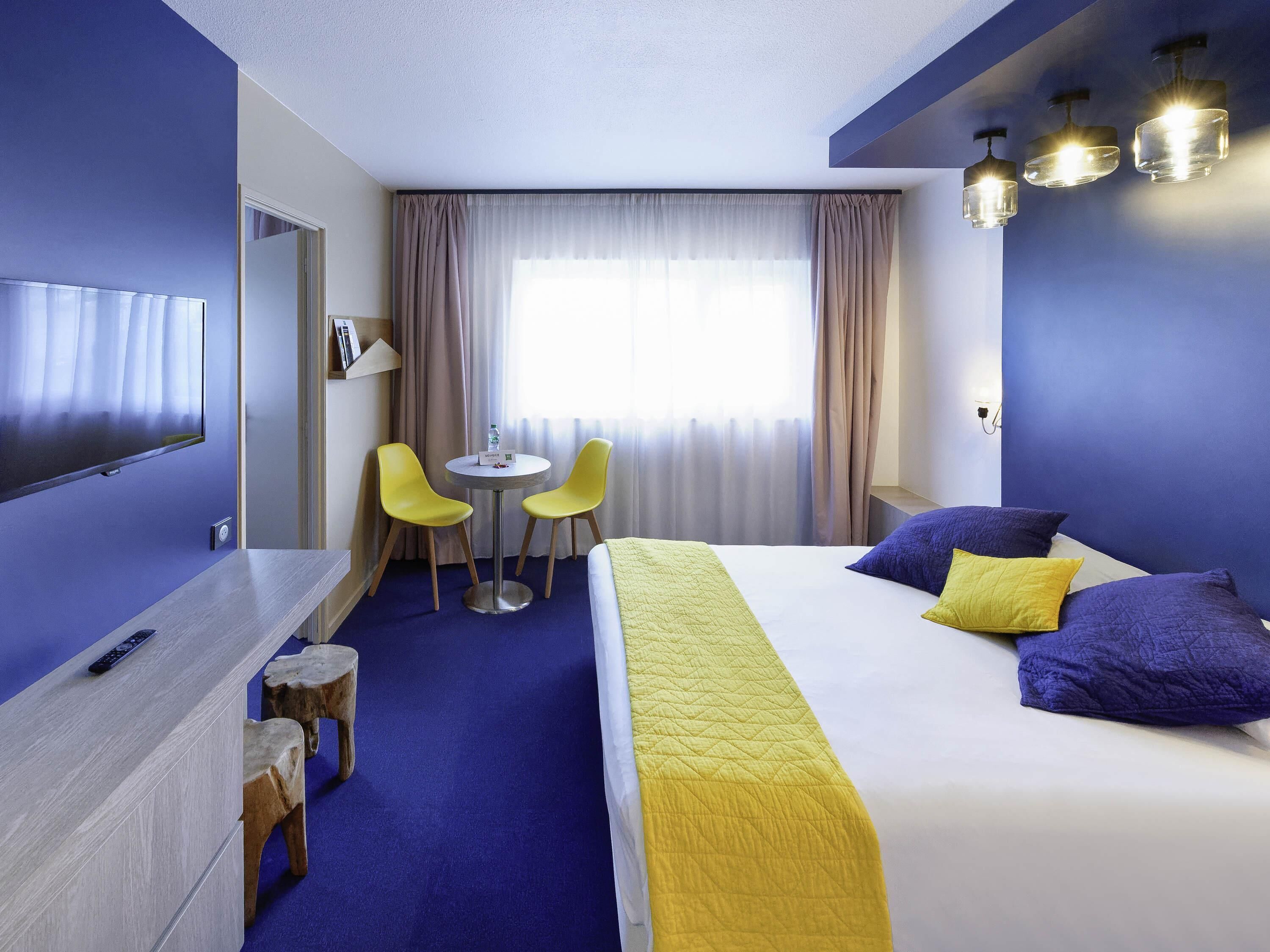 Ein Hotelzimmer mit Bett, Schreibtisch, zwei Stühlen, Fernseher und einem fensterbepflanzten Fenster.