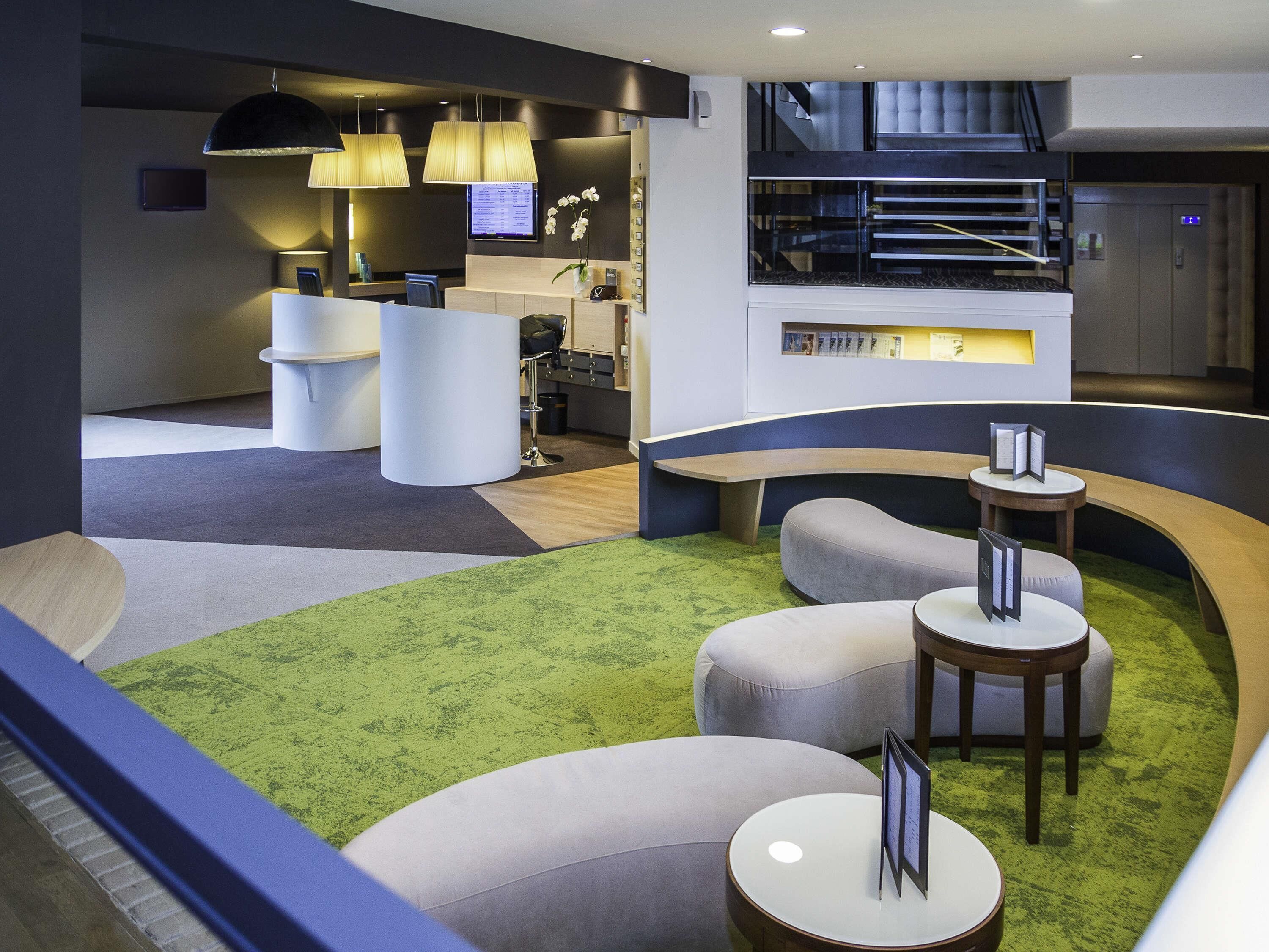 Un hall d’hôtel moderne comprenant un comptoir d’accueil, des espaces de détente et un escalier.
