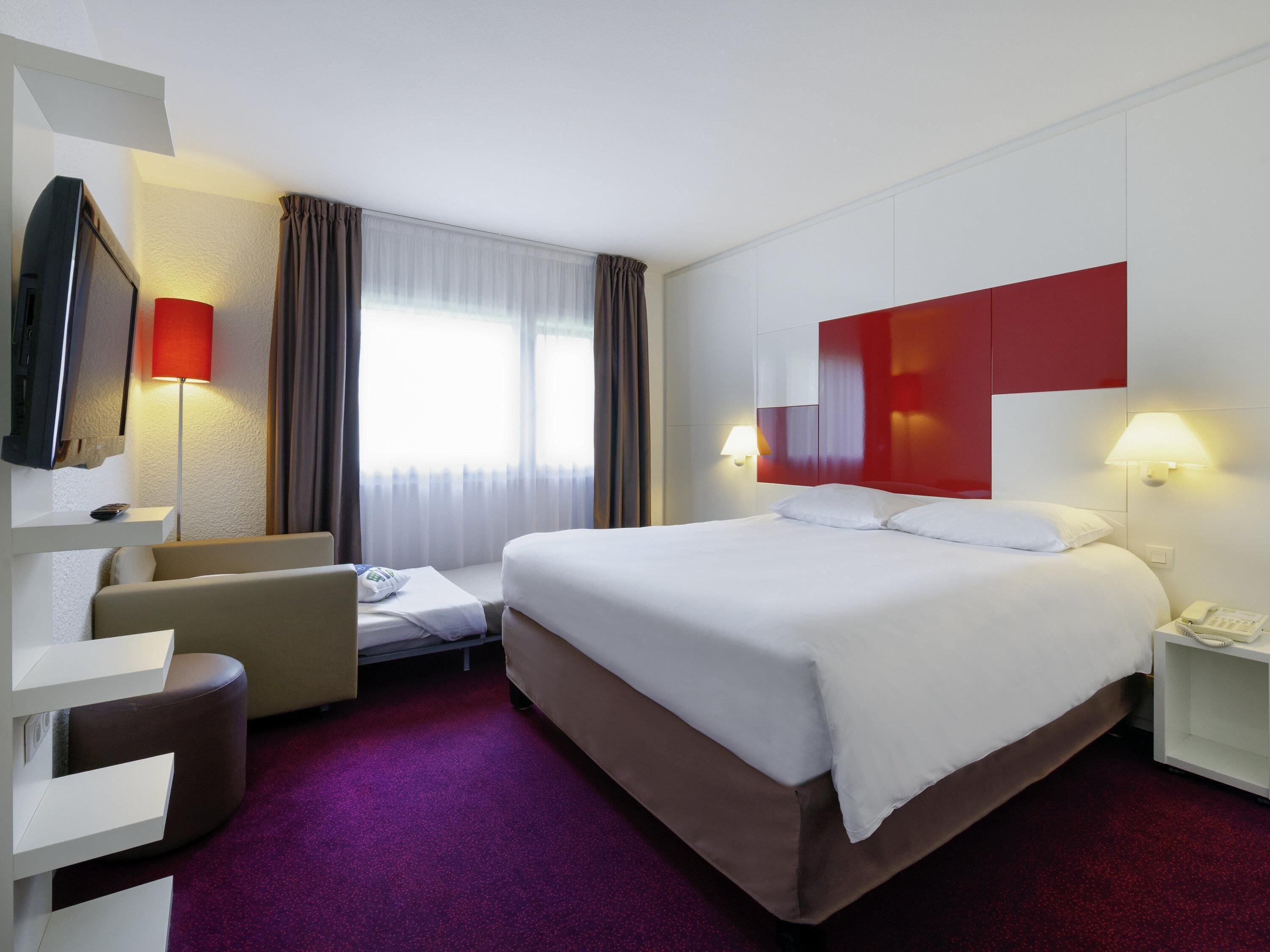 A hotel room with a bed, a desk, a chair, a TV, and a window with curtains.
