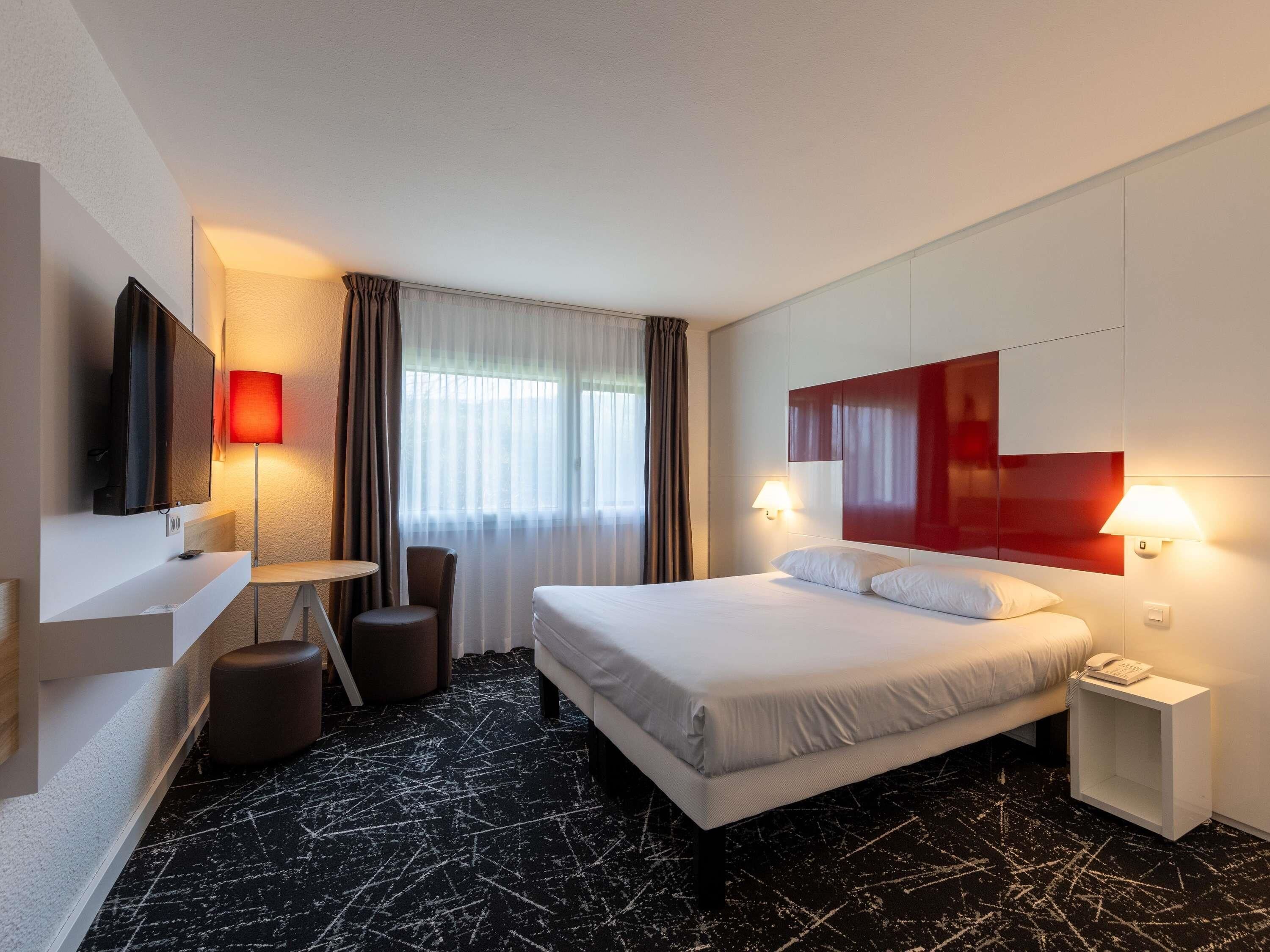 Habitación de hotel con cama, mesitas de noche, escritorio con silla, televisión y ventana con cortinas.