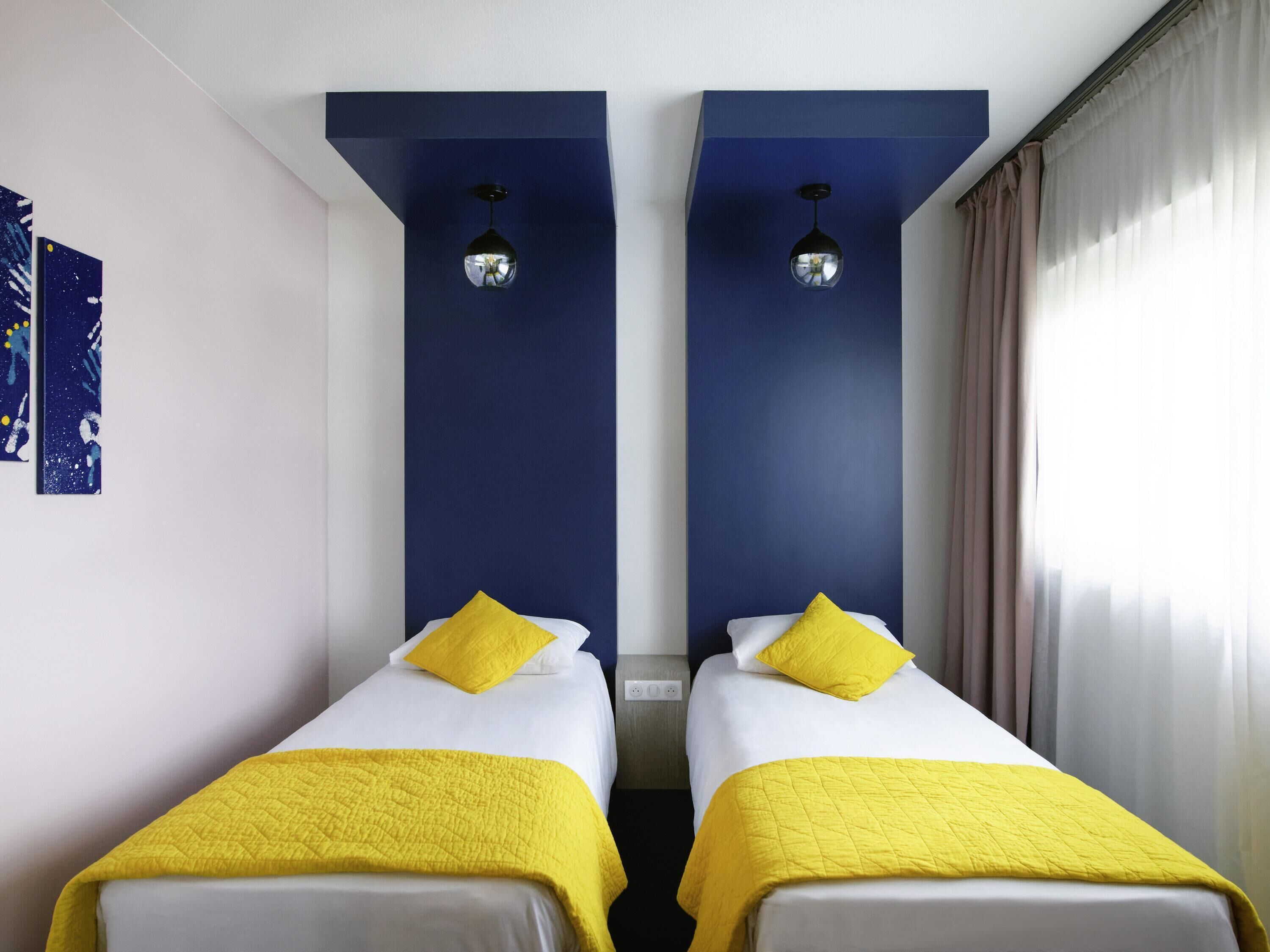 Ein Doppelzimmer mit zwei Betten, jedes mit einem gelben Kopfkissen und einer Decke, einer blauen Akzentwand und einer Deckenleuchte.