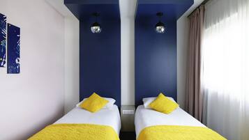 Ein Doppelzimmer mit zwei Betten, jedes mit einem gelben Kopfkissen und einer Decke, einer blauen Akzentwand und einer Deckenleuchte.