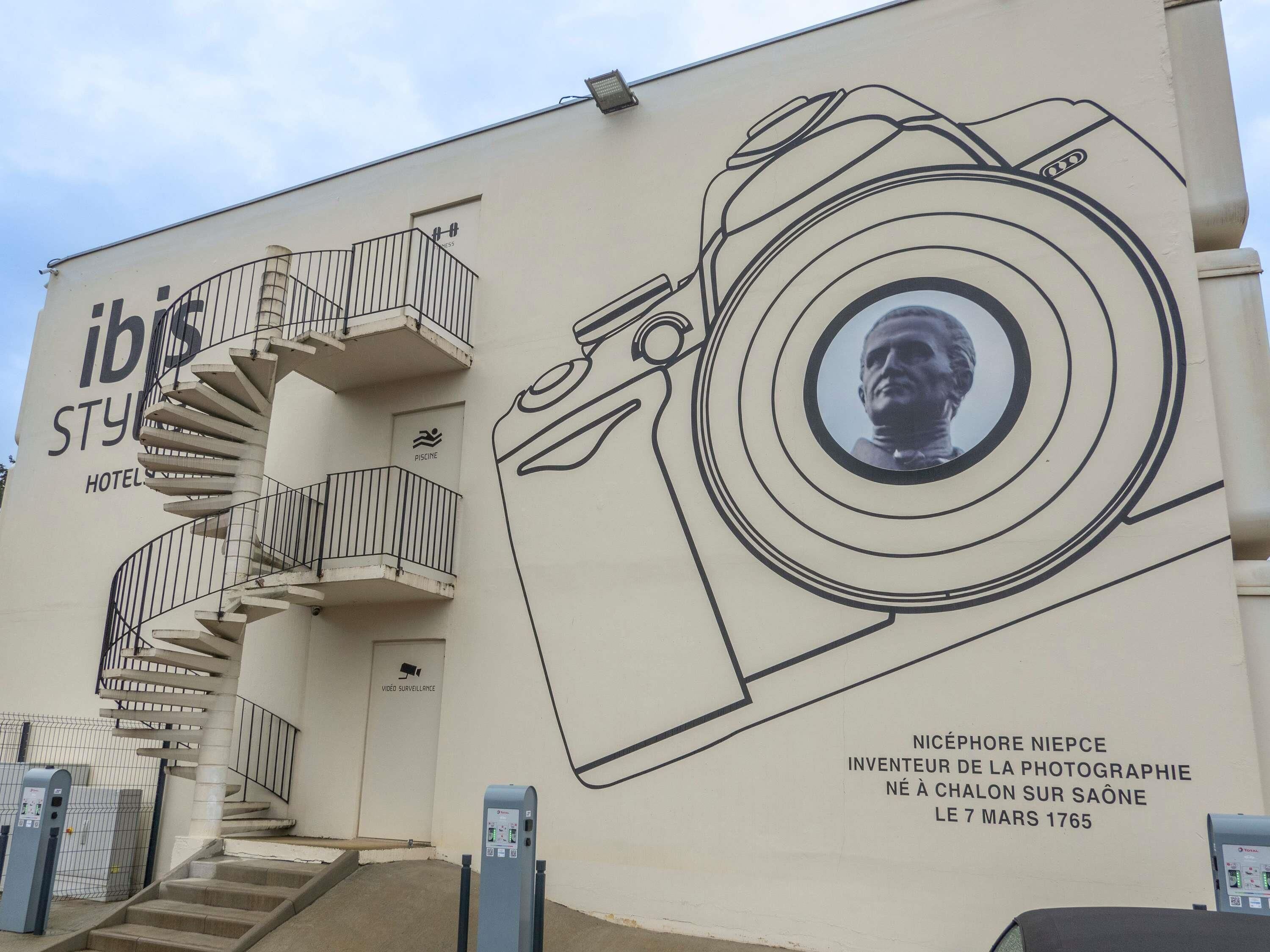 Un hôtel appelé « ibis » qui se distingue par un escalier en colimaçon, une fresque murale représentant un appareil photo et une plaque commémorative à la mémoire de Nicéphore Niépce.