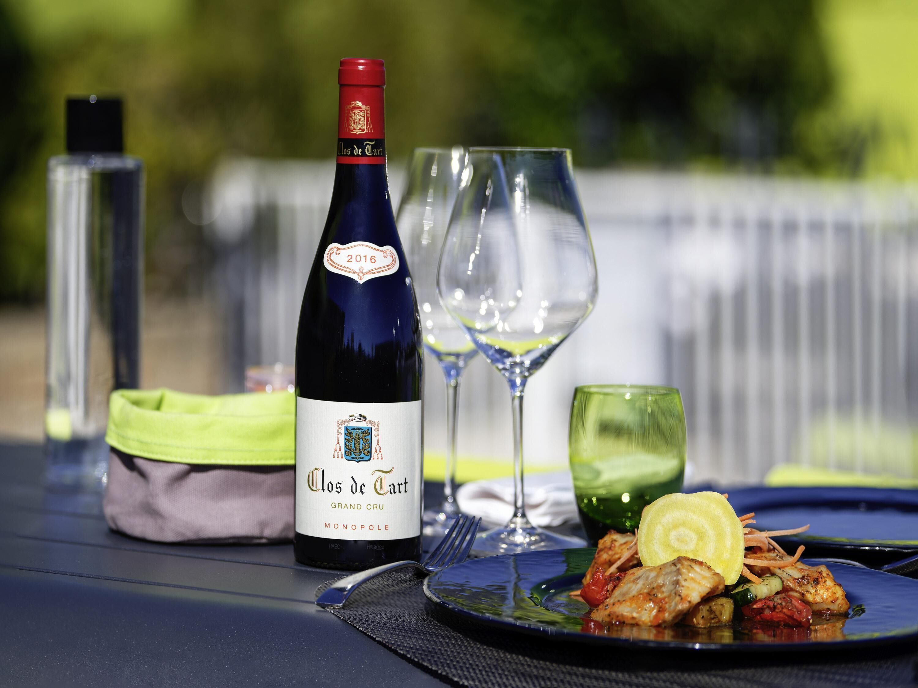 A bottle of Clos de Tart wine, a glass of water, and a plate of food on a table outdoors.