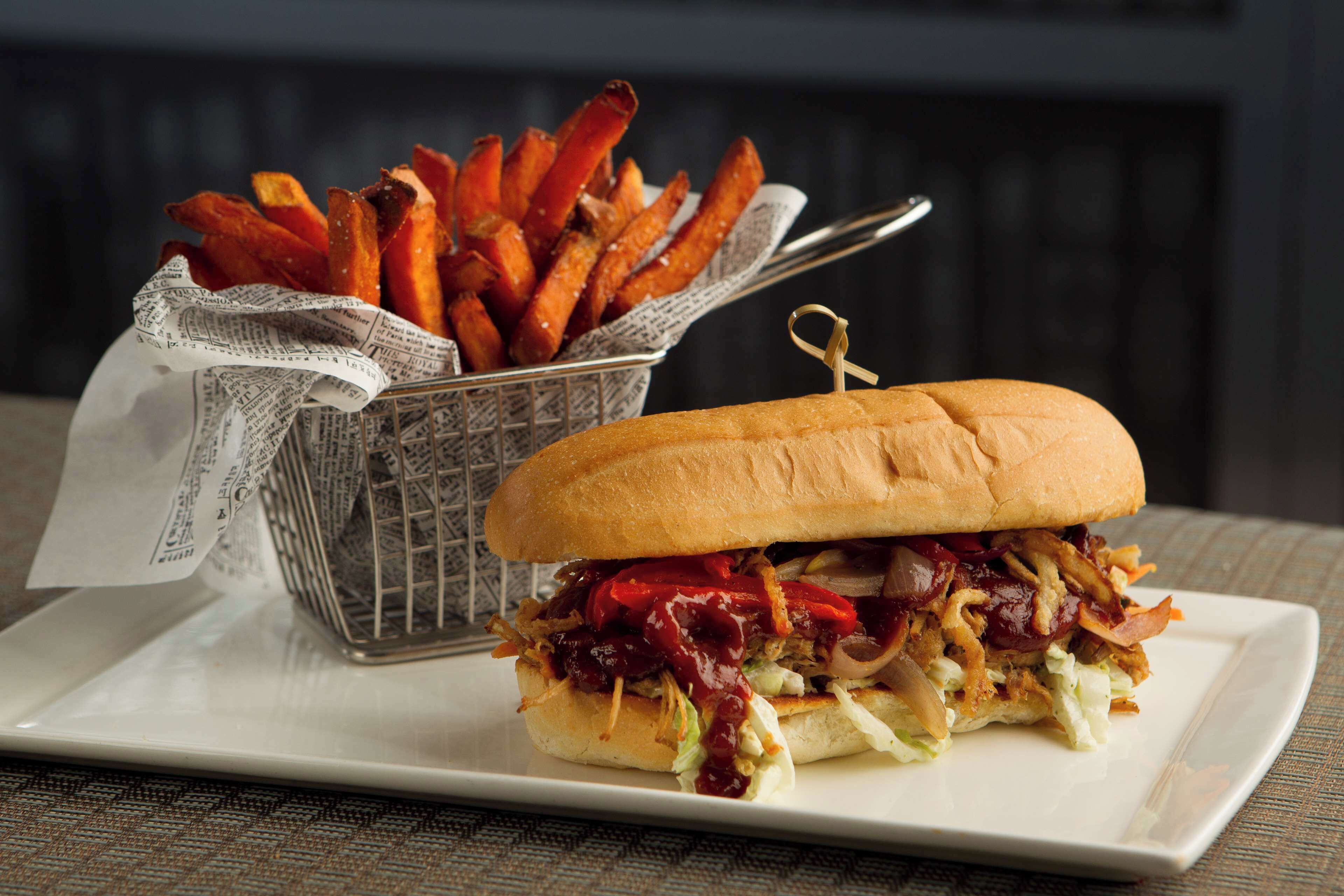 A sandwich with barbecue sauce and a side of sweet potato fries on a white plate.